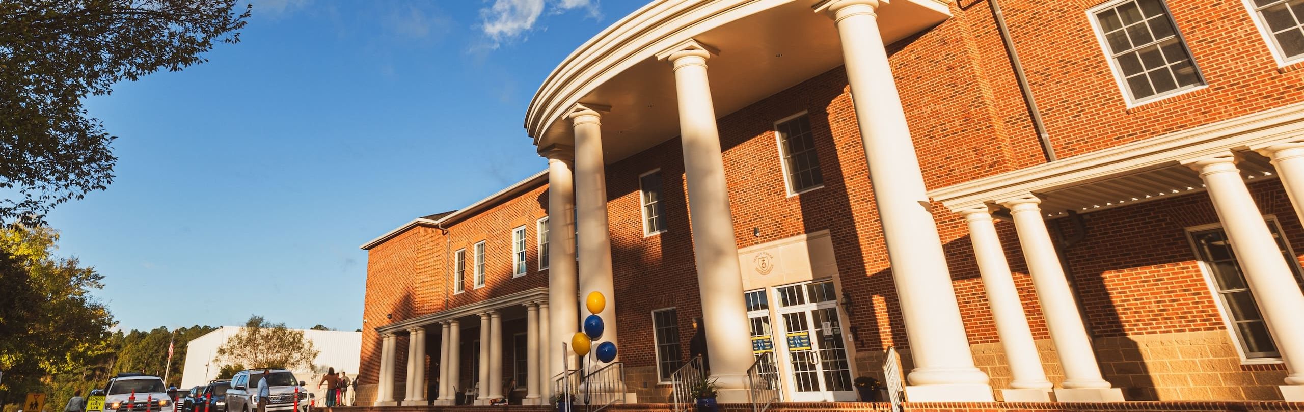  A warm, sunlit view of a classic red-brick school building with white columns and a portico, adorned with blue and yellow balloons. A clear sky with wispy clouds above.